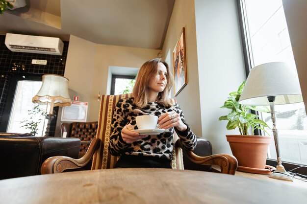 Una joven caucásica sentada en la mesa de un café cerca de la ventana, sosteniendo una taza de café o té en sus manos