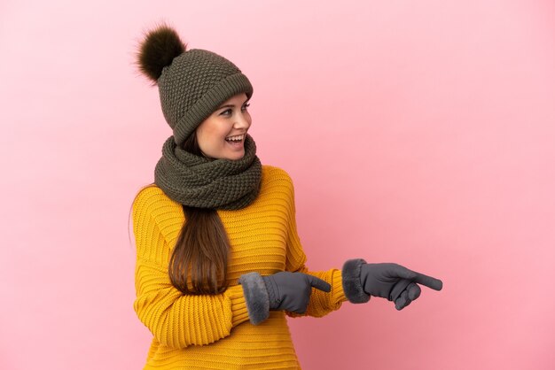 Foto joven caucásica con sombrero de invierno aislado sobre fondo rosa apuntando con el dedo hacia un lado y presentando un producto