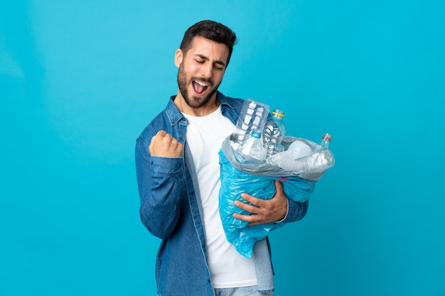 Joven caucásica sosteniendo una bolsa llena de botellas de plástico para reciclar aislado en la pared azul celebrando una victoria