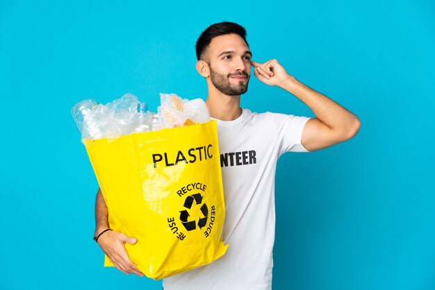 Joven caucásica sosteniendo una bolsa llena de botellas de plástico para reciclar aislado sobre fondo azul teniendo dudas y pensando