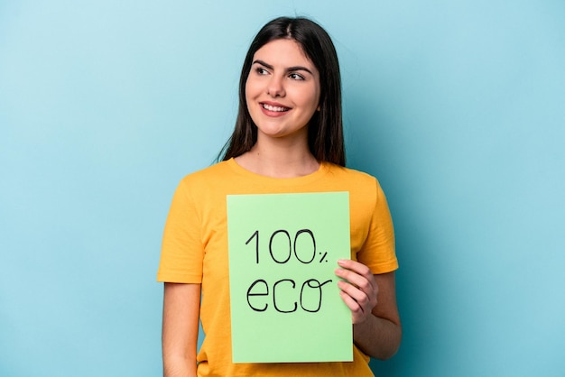 Una joven caucásica sosteniendo un cartel ecológico 100 aislado de fondo azul mira a un lado sonriendo alegre y agradable