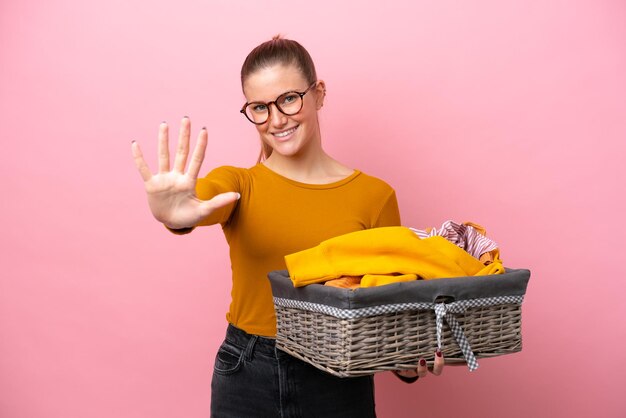 Joven caucásica sosteniendo una cesta de ropa aislada de fondo rosa contando cinco con los dedos