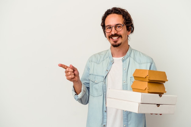 Joven caucásica sosteniendo una hamburguesa pizzas sonriendo y apuntando a un lado, mostrando algo