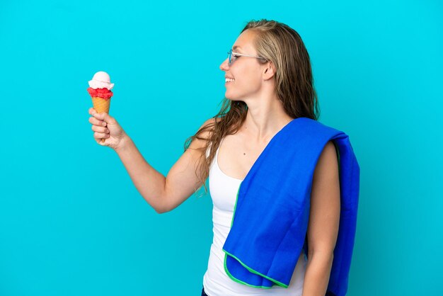 Joven caucásica sosteniendo helado y usando una toalla de playa aislada de fondo azul con expresión feliz