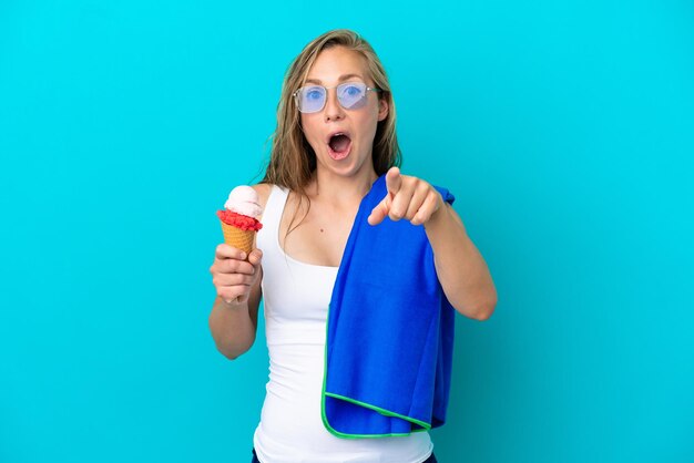 Joven caucásica sosteniendo helado y usando una toalla de playa aislada de fondo azul sorprendida y apuntando al frente