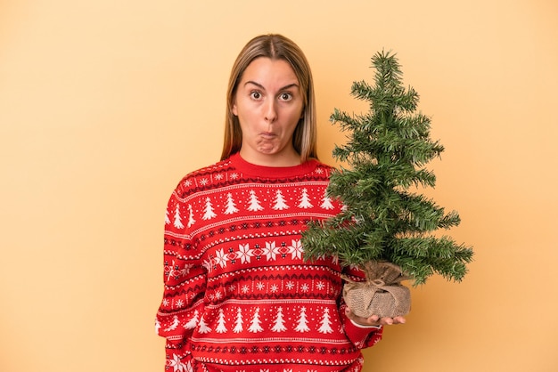 Una joven caucásica sosteniendo un pequeño árbol de navidad aislado en un fondo amarillo se encoge de hombros y abre los ojos confundidos.