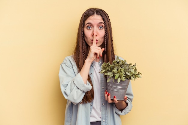 Joven caucásica sosteniendo una planta aislada de fondo amarillo guardando un secreto o pidiendo silencio.