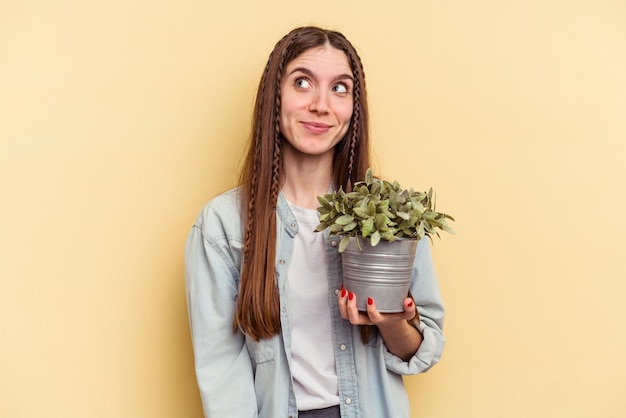 Joven caucásica sosteniendo una planta aislada de fondo amarillo soñando con lograr objetivos y propósitos