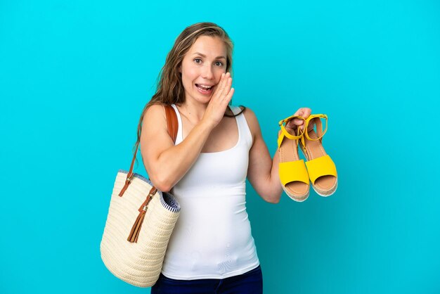 Joven caucásica sosteniendo sandalias de verano aisladas de fondo azul susurrando algo