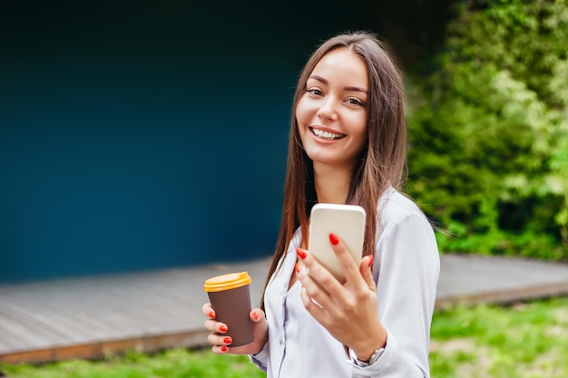 Una joven caucásica usa su teléfono inteligente sostiene una taza de café de papel y sonríe en el espacio de copia de la calle