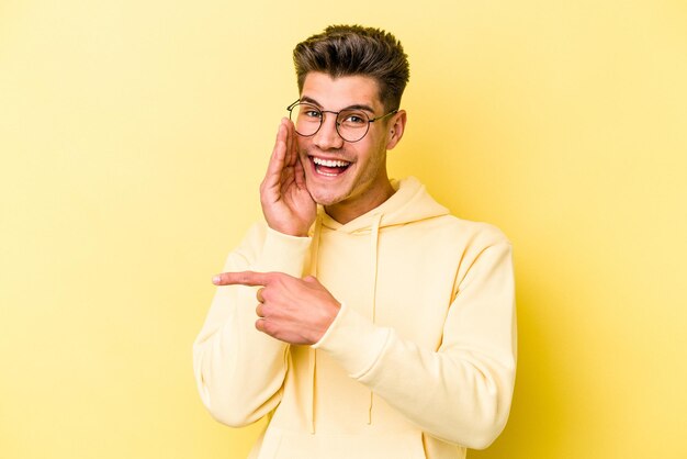 Joven caucásico aislado de fondo amarillo diciendo un chisme, señalando al lado informando algo.