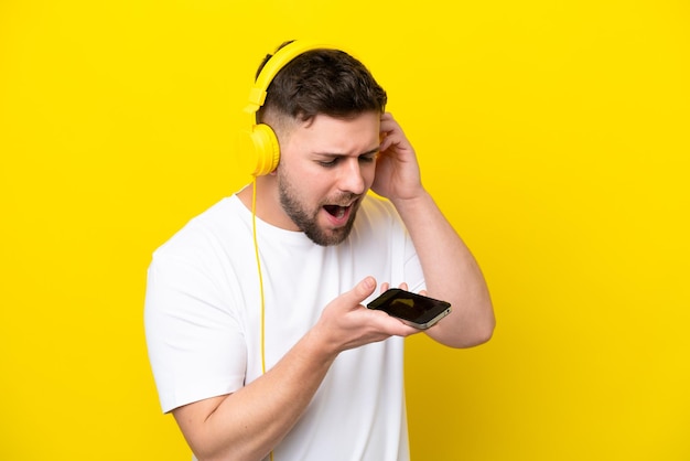 Joven caucásico aislado de fondo amarillo escuchando música con un móvil y cantando