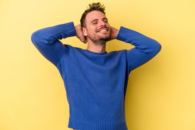 Joven caucásico aislado de fondo amarillo sintiéndose confiado, con las manos detrás de la cabeza.