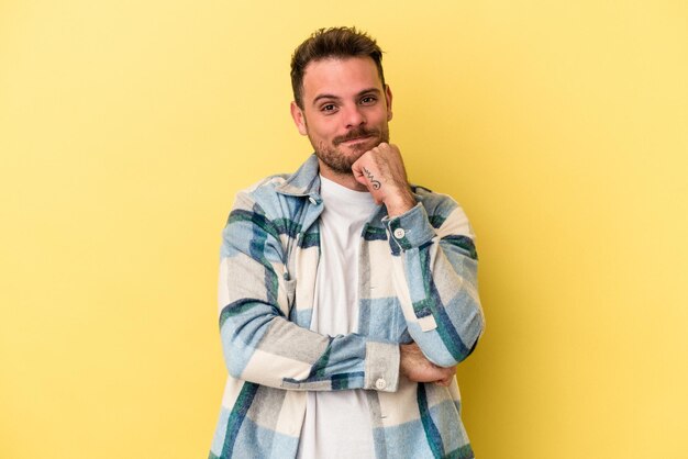 Joven caucásico aislado de fondo amarillo sonriendo feliz y confiado, tocando la barbilla con la mano.