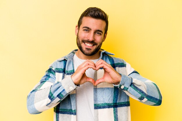 Joven caucásico aislado de fondo amarillo sonriendo y mostrando una forma de corazón con las manos