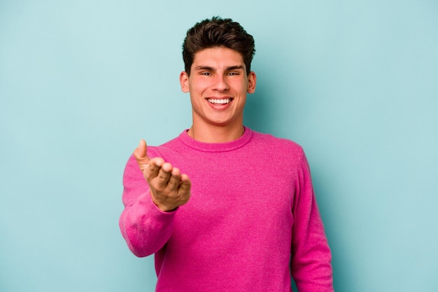 Joven caucásico aislado de fondo azul estirando la mano a la cámara en gesto de saludo