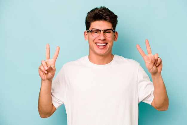 Joven caucásico aislado de fondo azul que muestra el signo de la victoria y sonríe ampliamente