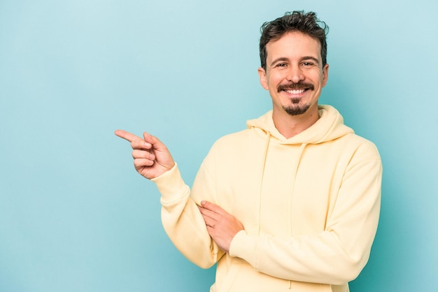 Joven caucásico aislado de fondo azul sonriendo alegremente señalando con el dedo índice.