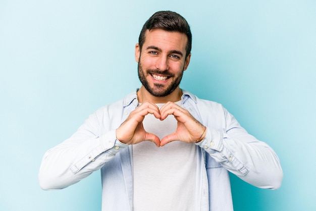 Joven caucásico aislado de fondo azul sonriendo y mostrando una forma de corazón con las manos