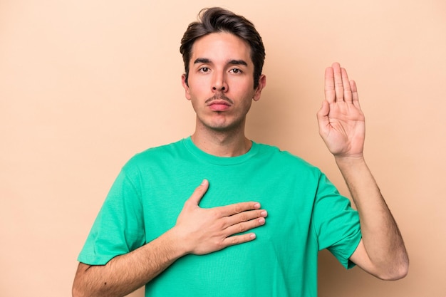 Joven caucásico aislado de fondo beige haciendo un juramento, poniendo la mano en el pecho.