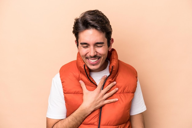 Joven caucásico aislado de fondo beige se ríe a carcajadas manteniendo la mano en el pecho