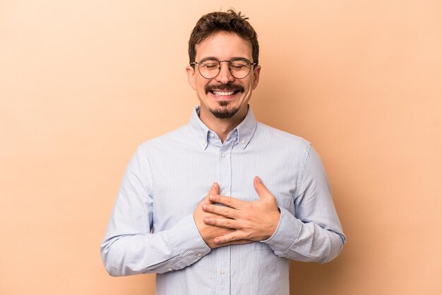 Joven caucásico aislado de fondo beige riéndose manteniendo las manos en el corazón, concepto de felicidad.