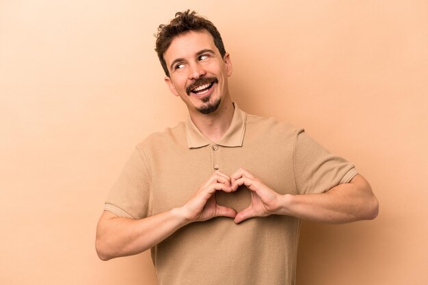 Joven caucásico aislado de fondo beige sonriendo y mostrando una forma de corazón con las manos