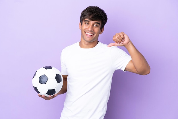 Joven caucásico aislado de fondo morado con balón de fútbol y orgulloso de sí mismo