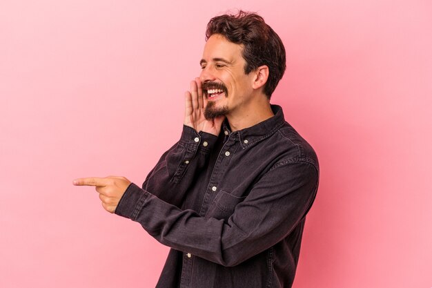 Joven caucásico aislado de fondo rosa diciendo un chisme, señalando al lado informando algo.