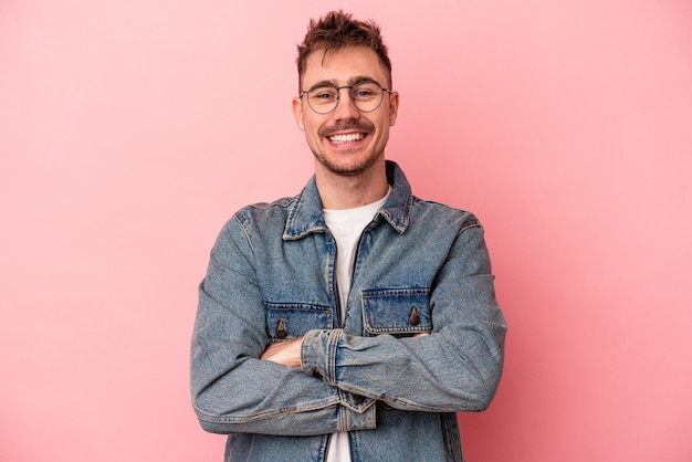 Joven caucásico aislado de fondo rosa que se siente confiado, cruzando los brazos con determinación.