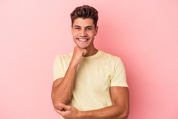 Joven caucásico aislado de fondo rosa sonriendo feliz y confiado, tocando la barbilla con la mano.