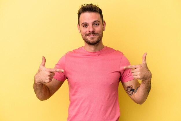 Joven caucásico aislado en una persona de fondo amarillo que señala con la mano un espacio de copia de camisa, orgulloso y confiado