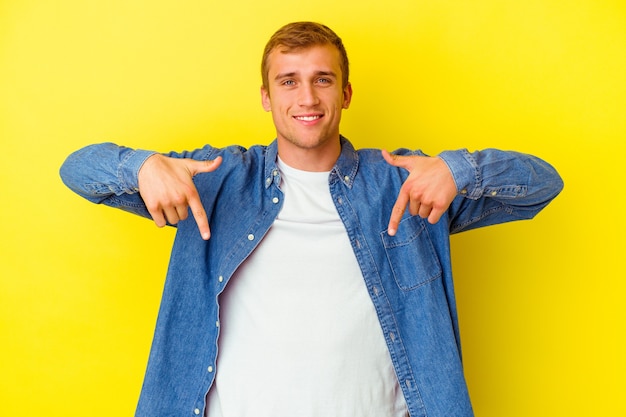 Joven caucásico aislado sobre fondo amarillo apunta hacia abajo con los dedos, sentimiento positivo.