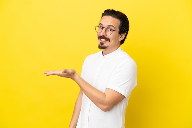 Joven caucásico aislado sobre fondo amarillo presentando una idea mientras mira sonriendo hacia