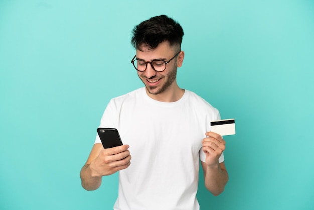 Joven caucásico aislado sobre fondo azul comprando con el móvil con tarjeta de crédito