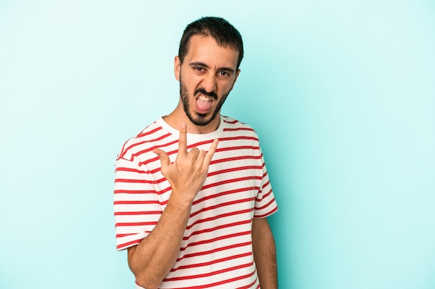 Joven caucásico aislado sobre fondo azul mostrando gesto de rock con los dedos