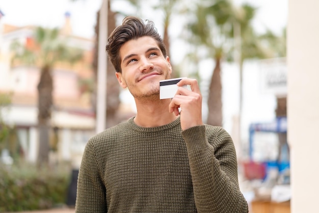 Joven caucásico al aire libre sosteniendo una tarjeta de crédito y pensando