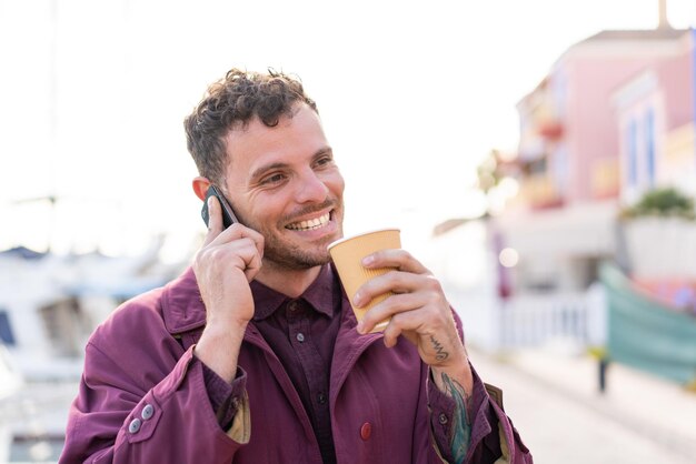 Joven caucásico al aire libre usando un teléfono móvil y sosteniendo un café con expresión feliz