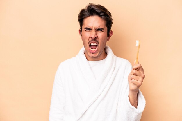 Joven caucásico con albornoz sosteniendo un cepillo de dientes aislado de fondo beige gritando muy enojado y agresivo