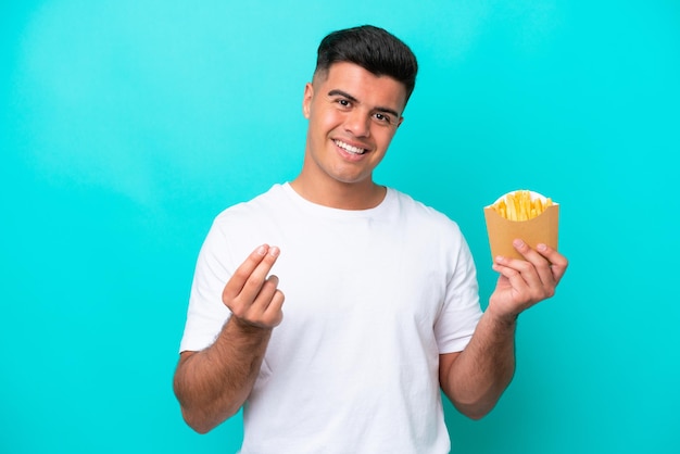 Joven caucásico atrapando papas fritas aisladas de fondo azul haciendo gestos de dinero