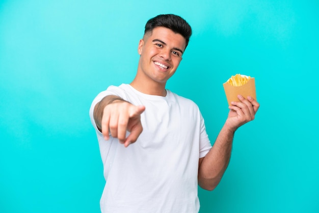 Joven caucásico atrapando papas fritas aislado en fondo azul apuntando al frente con expresión feliz