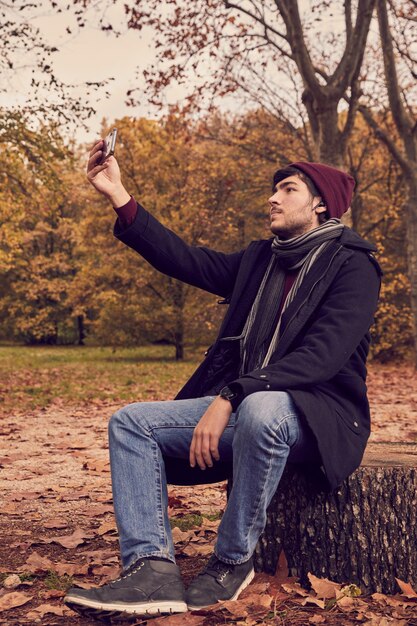 Joven caucásico blanco sentado en un tronco tomando un selfie con un teléfono inteligente con auriculares bluetooth y un reloj inteligente en el bosque Colores de otoño