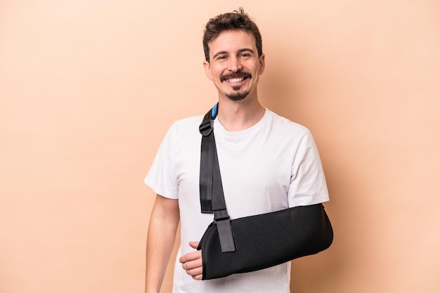 Joven caucásico con el brazo roto aislado de fondo beige feliz, sonriente y alegre.