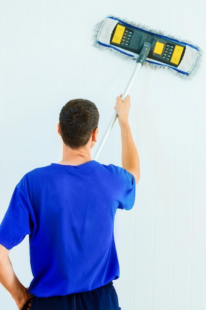 Un joven caucásico con una camiseta azul lava la pared blanca de la habitación con un trapeador