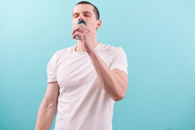 Joven caucásico con una camiseta blanca bebiendo una bebida energética con los ojos cerrados sobre fondo azul.