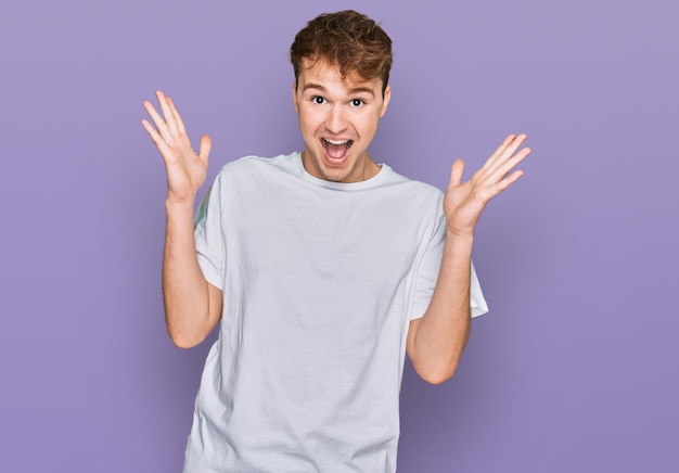 Joven caucásico con camiseta blanca casual celebrando loco y asombrado por el éxito con los brazos levantados y los ojos abiertos gritando emocionado concepto de ganador