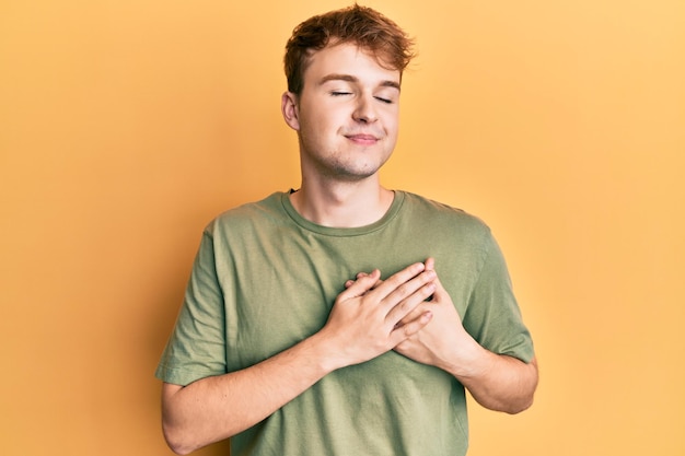 Joven caucásico con camiseta verde casual sonriendo con las manos en el pecho con los ojos cerrados con un gesto agradecido en el concepto de salud facial