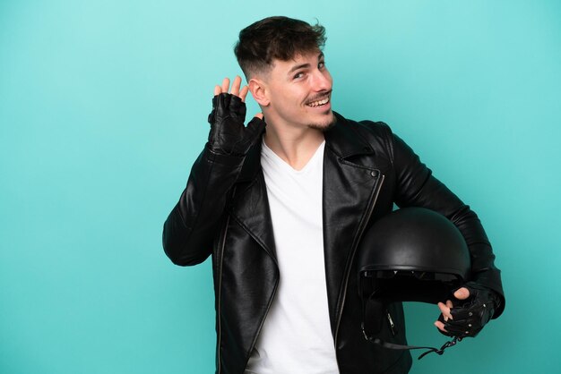 Joven caucásico con casco de moto aislado de fondo azul escuchando algo poniendo la mano en la oreja