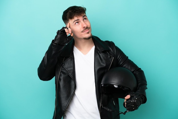 Joven caucásico con casco de motocicleta aislado de fondo azul con dudas y pensando