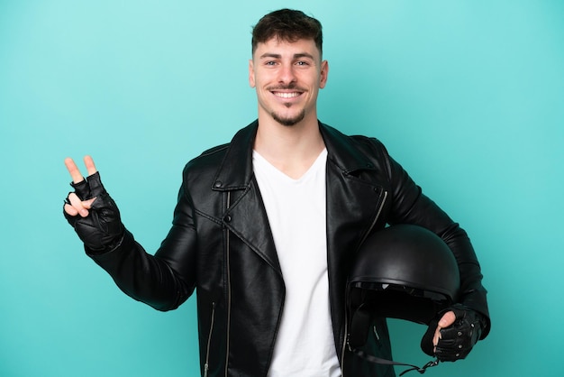 Joven caucásico con casco de motocicleta aislado de fondo azul sonriendo y mostrando el signo de la victoria